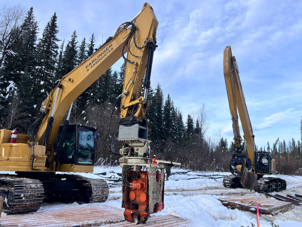 Two yellow excavators on a snowy construction site near pine trees, one equipped with a drilling attachment.