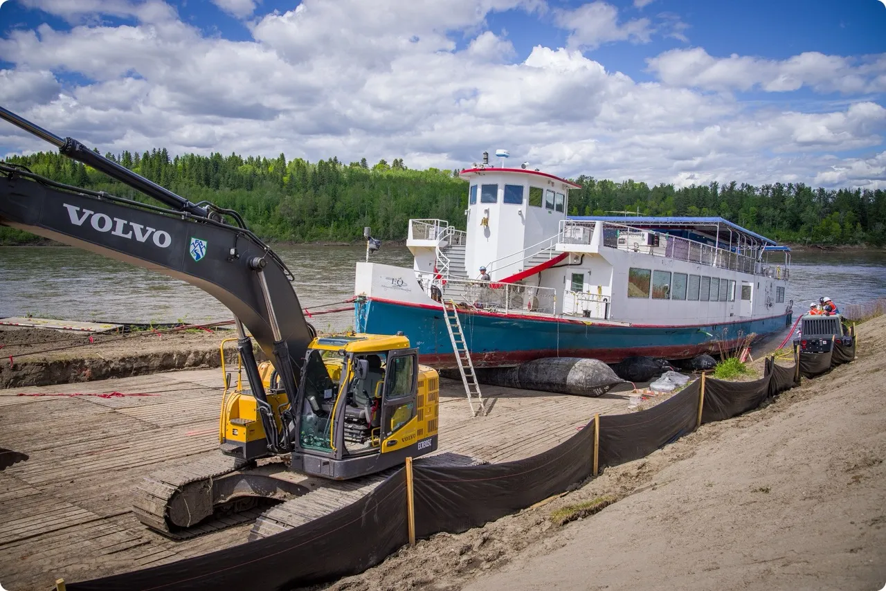 White and blue riverboat on a wooden platform near a river, next to a yellow Volvo excavator with a forested background under a partly cloudy sky.