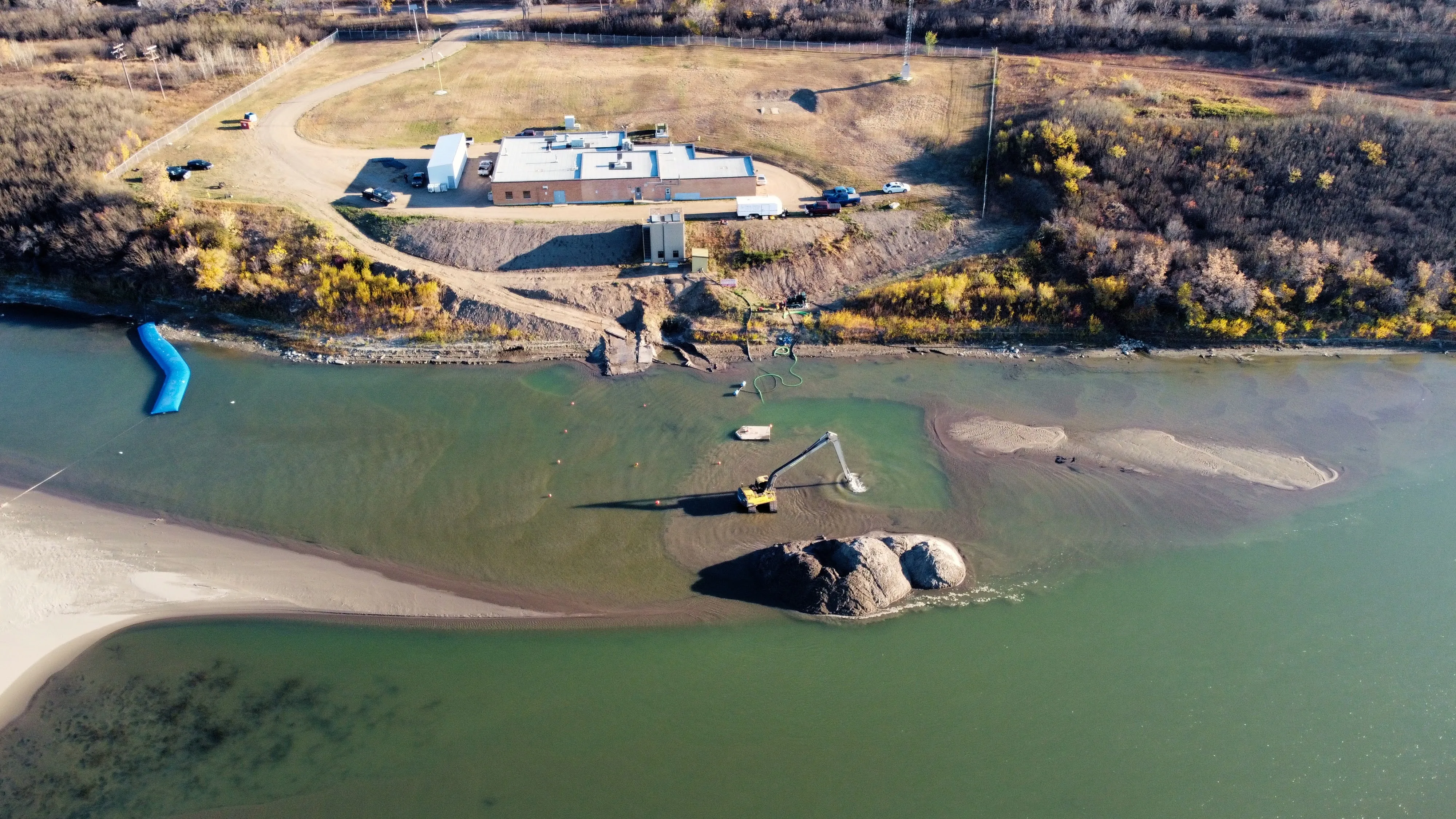 Aerial view of a riverbank restoration site with machinery dredging near sandbars and a building in the background.