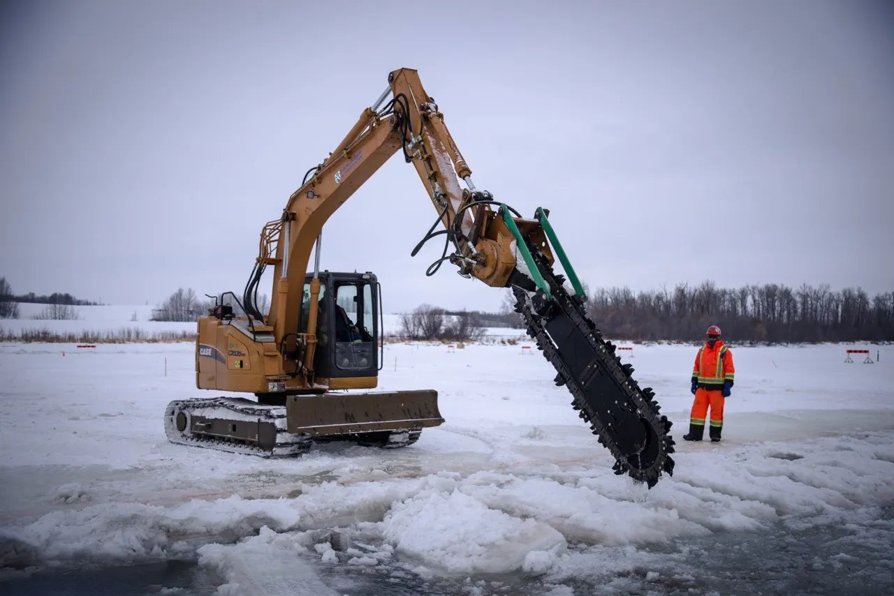 Heavy excavator with a large cutting attachment clearing snow and ice on a frozen landscape with a worker in orange safety gear nearby.
