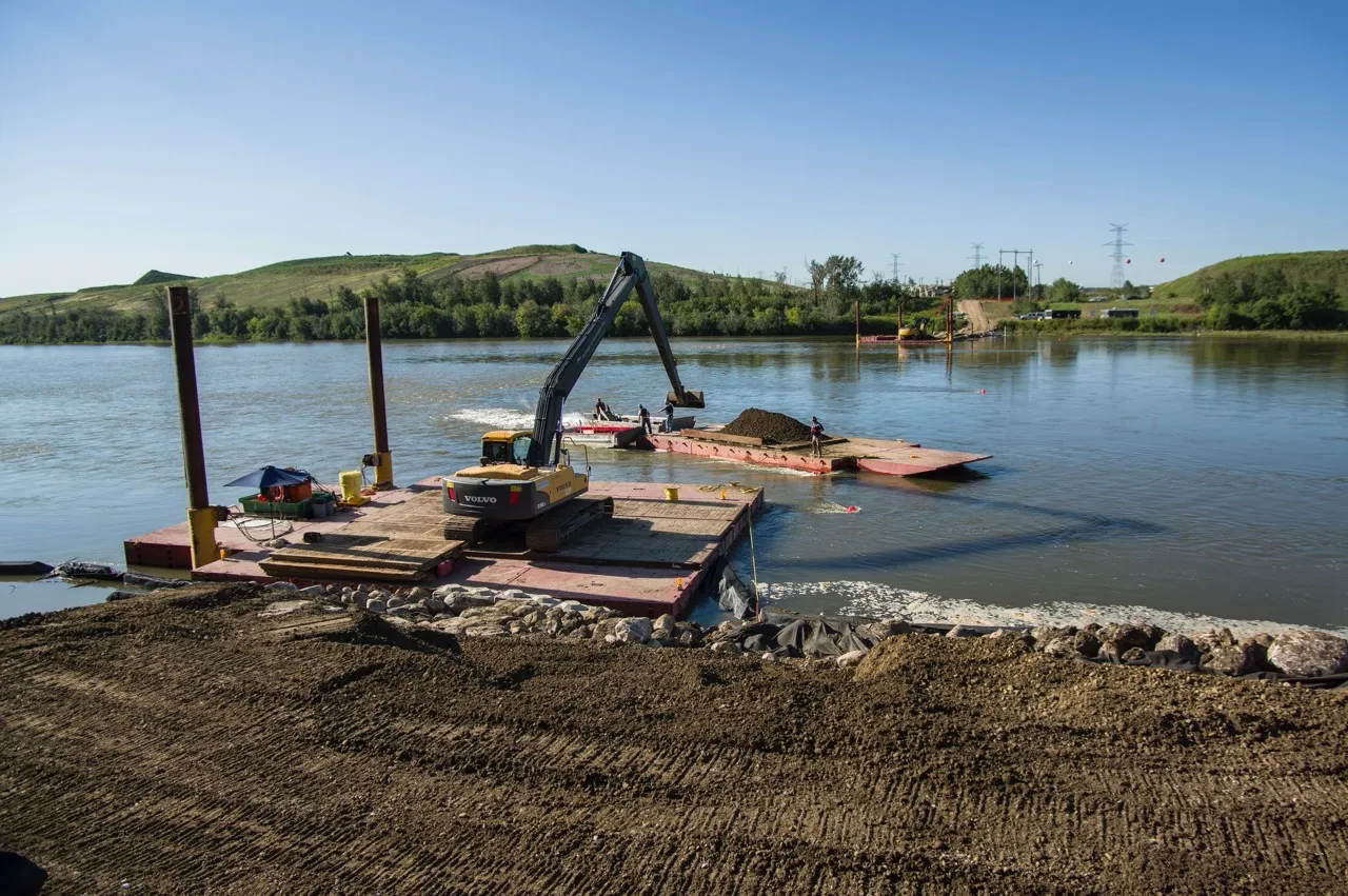 Excavator loading dirt onto a barge on a river, with hills and trees in the background under a clear blue sky.