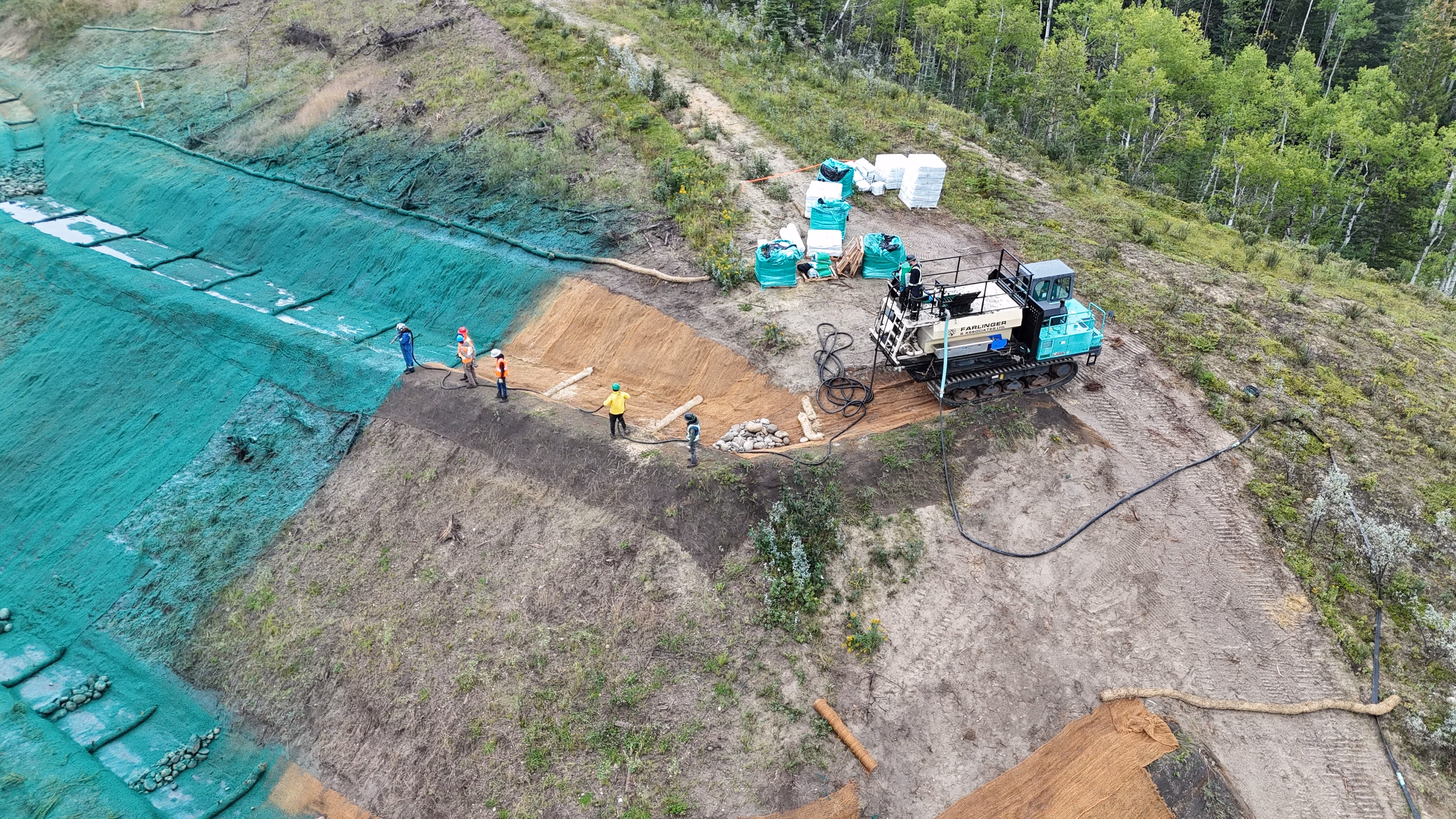 Yellow excavator working near a riverbank with workers and a wooden barrier in a forested area under a cloudy sky.