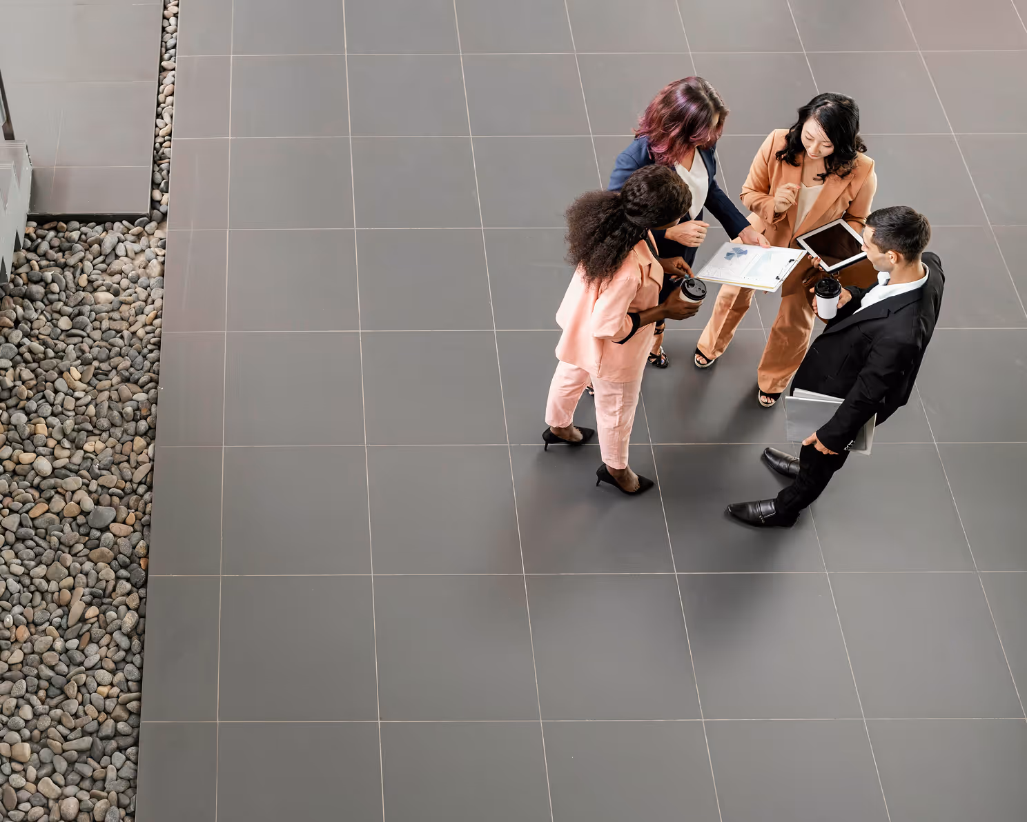 Overhead view of a diverse group of four business professionals standing on a tiled floor, discussing documents and a tablet.