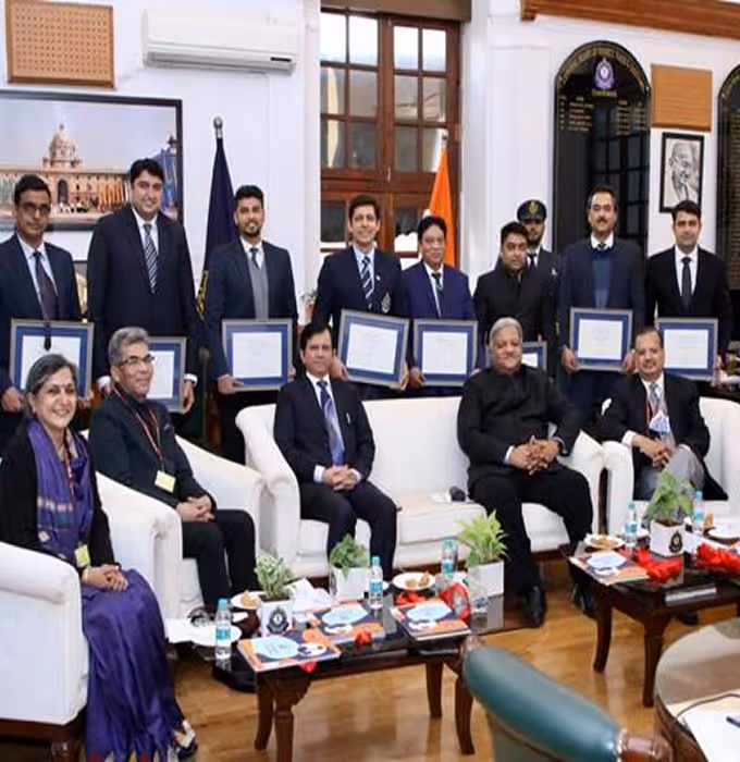 Group of men and women, some seated on white sofas and others standing behind holding certificates, in a formal room with framed photos and an Indian flag.