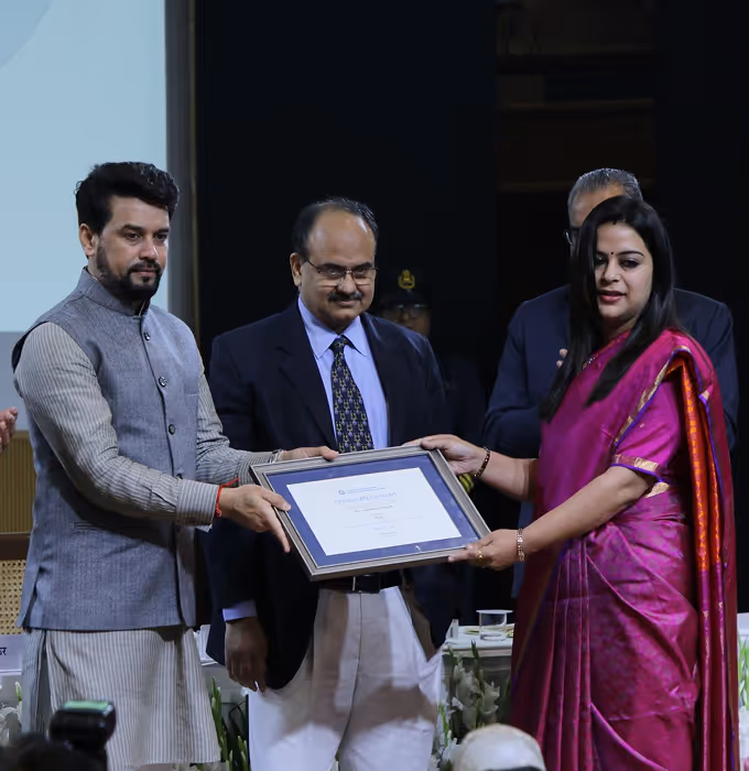 A woman in a pink saree and a man in traditional Indian attire hold a framed certificate during an indoor award ceremony with men standing in the background.