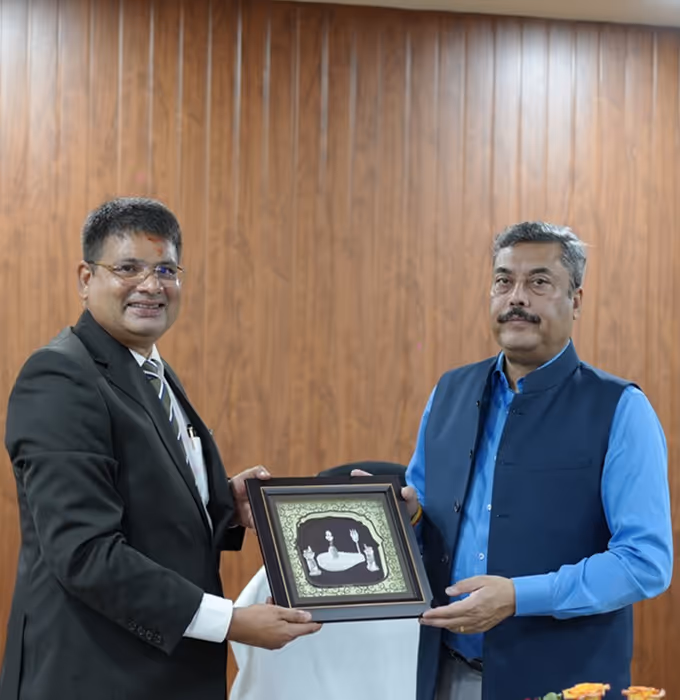 Two men in formal attire holding and presenting a framed decorative artwork together against a wooden paneled wall.