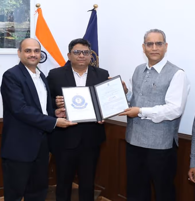 Three men in formal attire holding and displaying a certificate in a room with Indian and institutional flags in the background.