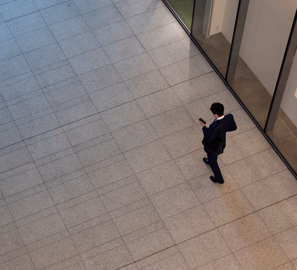 Businessman in a navy suit with a black tote bag standing on a tiled floor, looking at his smartphone near glass office walls.