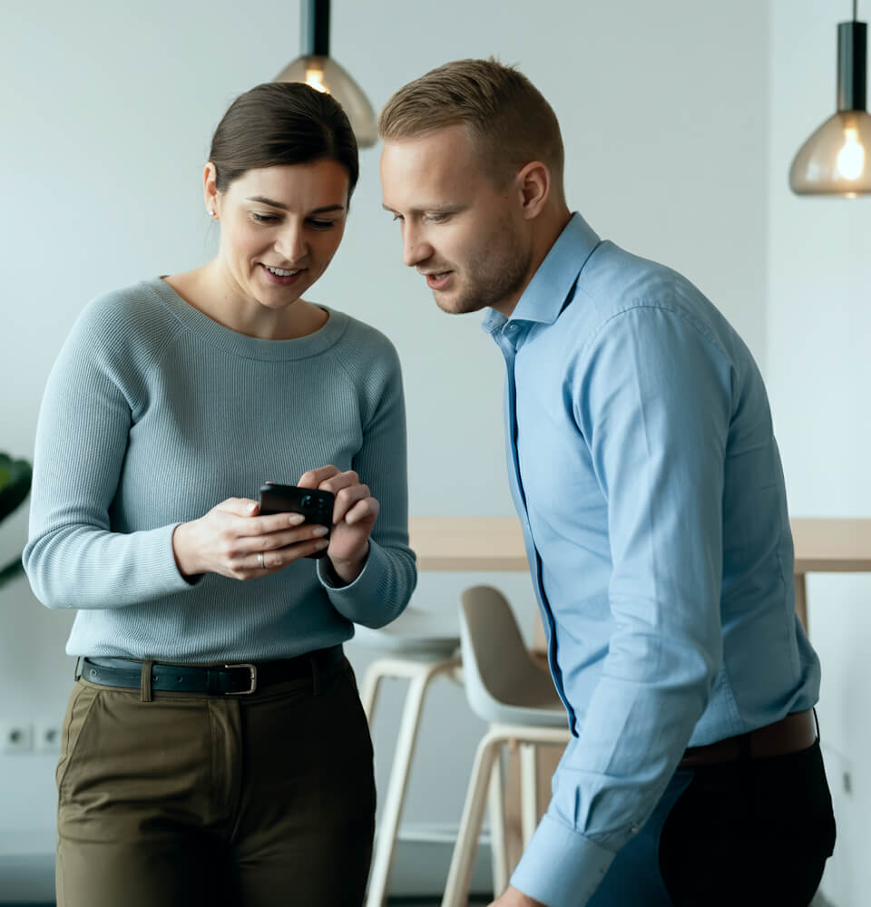 Two coworkers, a woman and a man, looking at a smartphone together in a modern office setting.