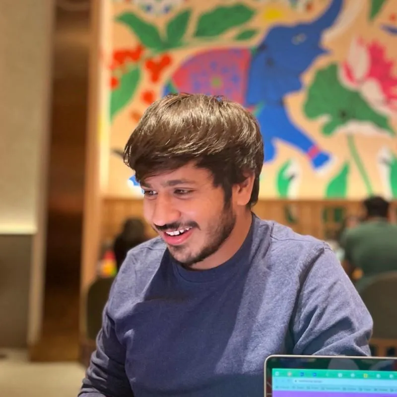 Smiling young man with dark hair and beard wearing a blue shirt, sitting indoors with a colorful mural featuring an elephant in the background and a laptop screen partially visible in front.