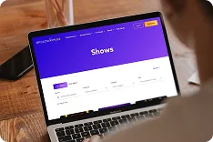Person looking at a laptop screen displaying a purple-themed website page titled 'Shows' with filter options and a table layout.