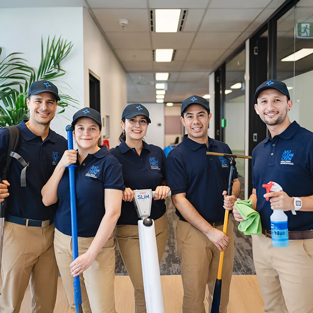 The Just Clean Crew staff are smiling, wearing navy polo shirts and caps, holding various cleaning tools and supplies in an office hallway.