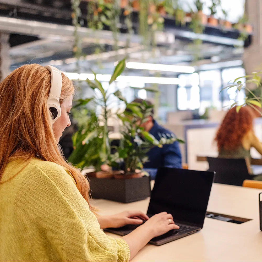 Woman in yellow sweater wearing headphones and typing on a laptop at a modern office desk with plants and coworkers in the background.