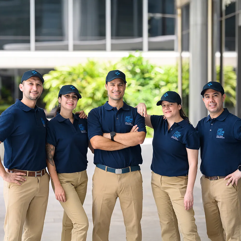 Five of the Just Clean Crew in navy blue polo shirts and caps with khaki pants are posing and smiling outdoors in a workplace setting.