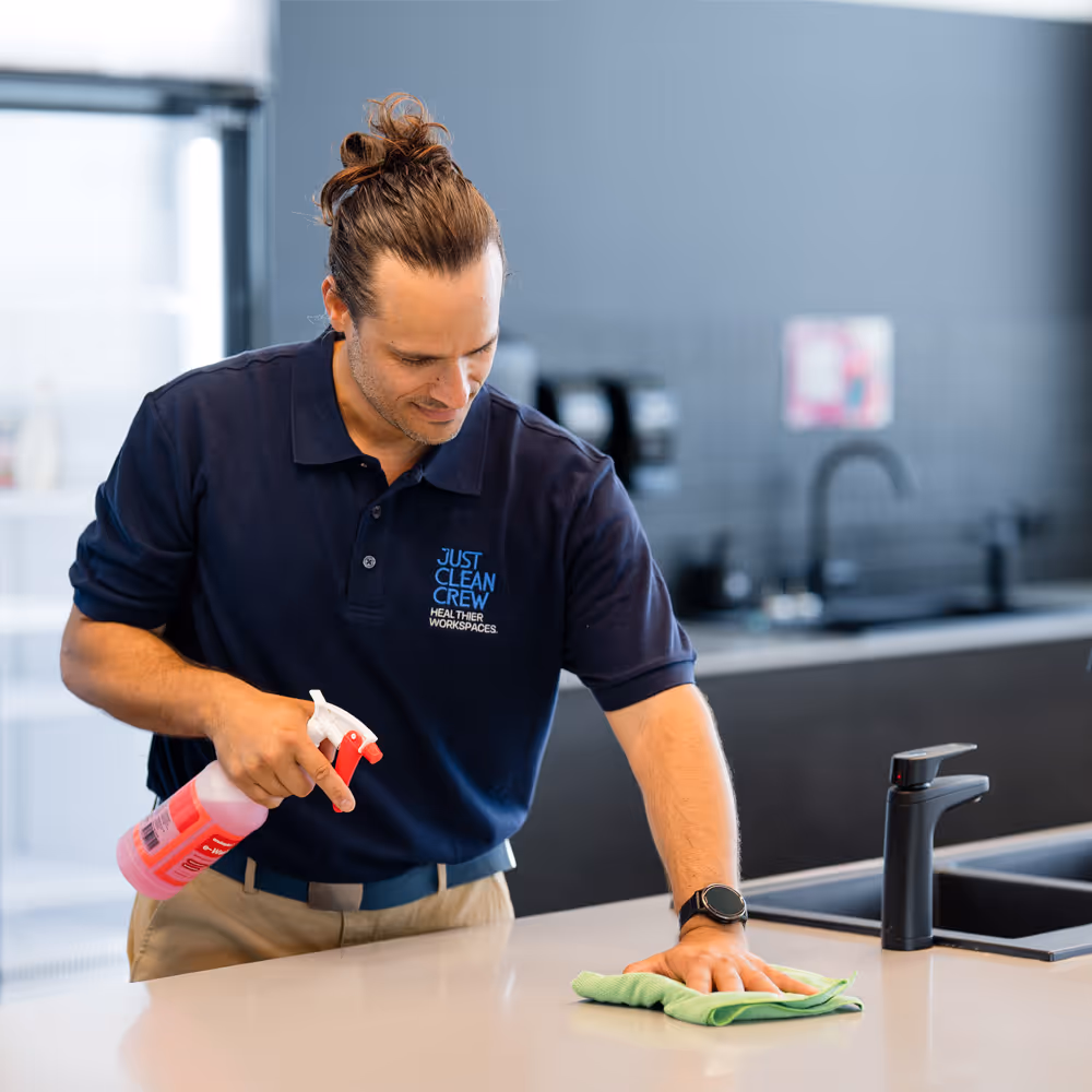 Just Clean Crew team member with tied-back hair wearing a navy shirt spraying and wiping a clean kitchen counter with a green cloth.