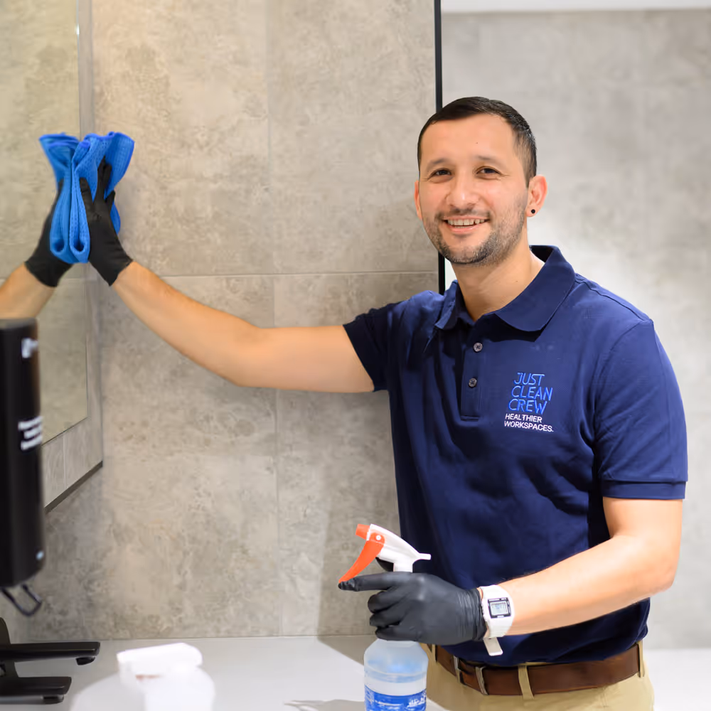 Smiling Just Clean Crew team member wearing black gloves and a blue Just Clean Crew shirt, wiping a mirror with a blue cloth and holding a spray bottle.