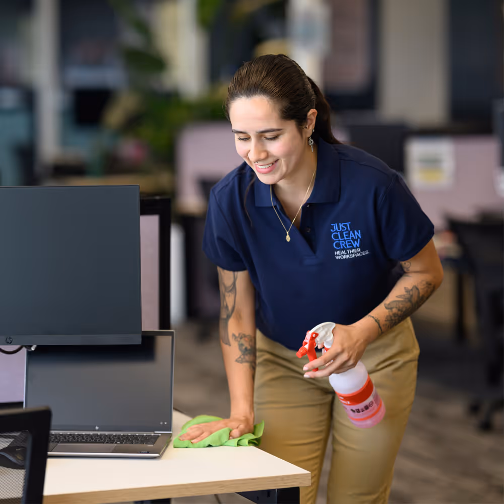 Smiling Just Clean Crew team member in a navy blue Just Clean Crew shirt, cleaning an office desk with a green cloth and a spray bottle.