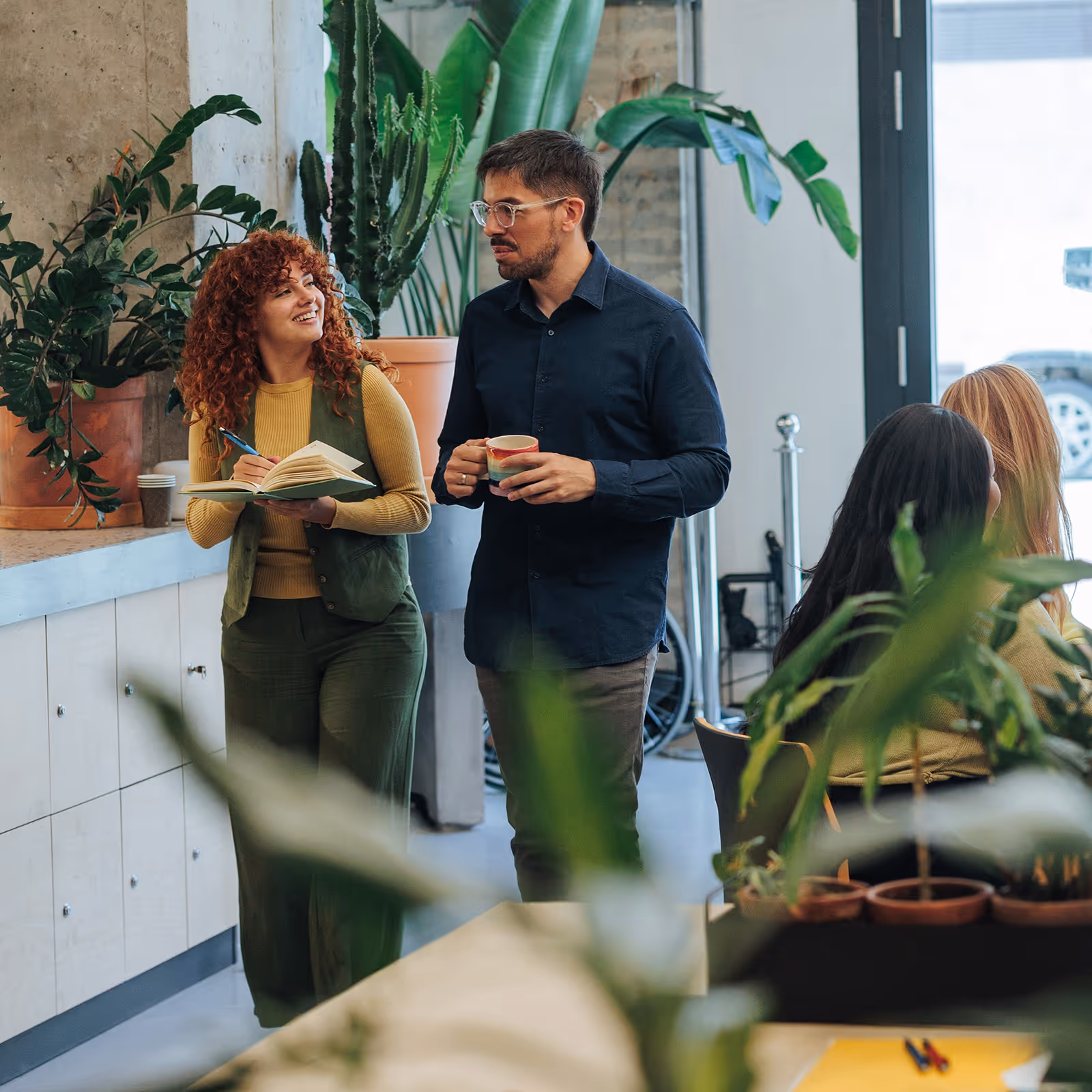 Two colleagues, a woman writing in a notebook and a man holding a coffee cup, smiling and chatting in a clean modern office with large green plants.
