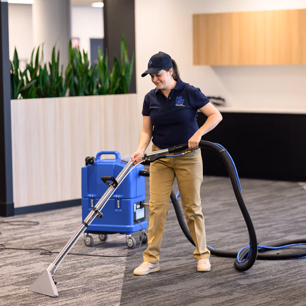 A Just Clean Crew team member cleans a carpet with a blue professional carpet cleaning machine in a modern office setting.