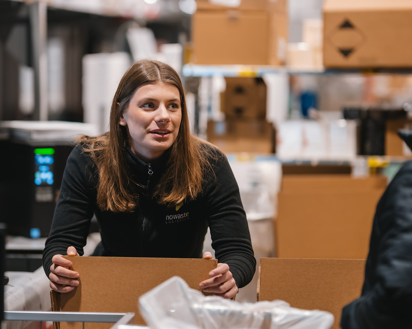 Woman in a black jacket packing a cardboard box in a warehouse filled with stacked boxes.