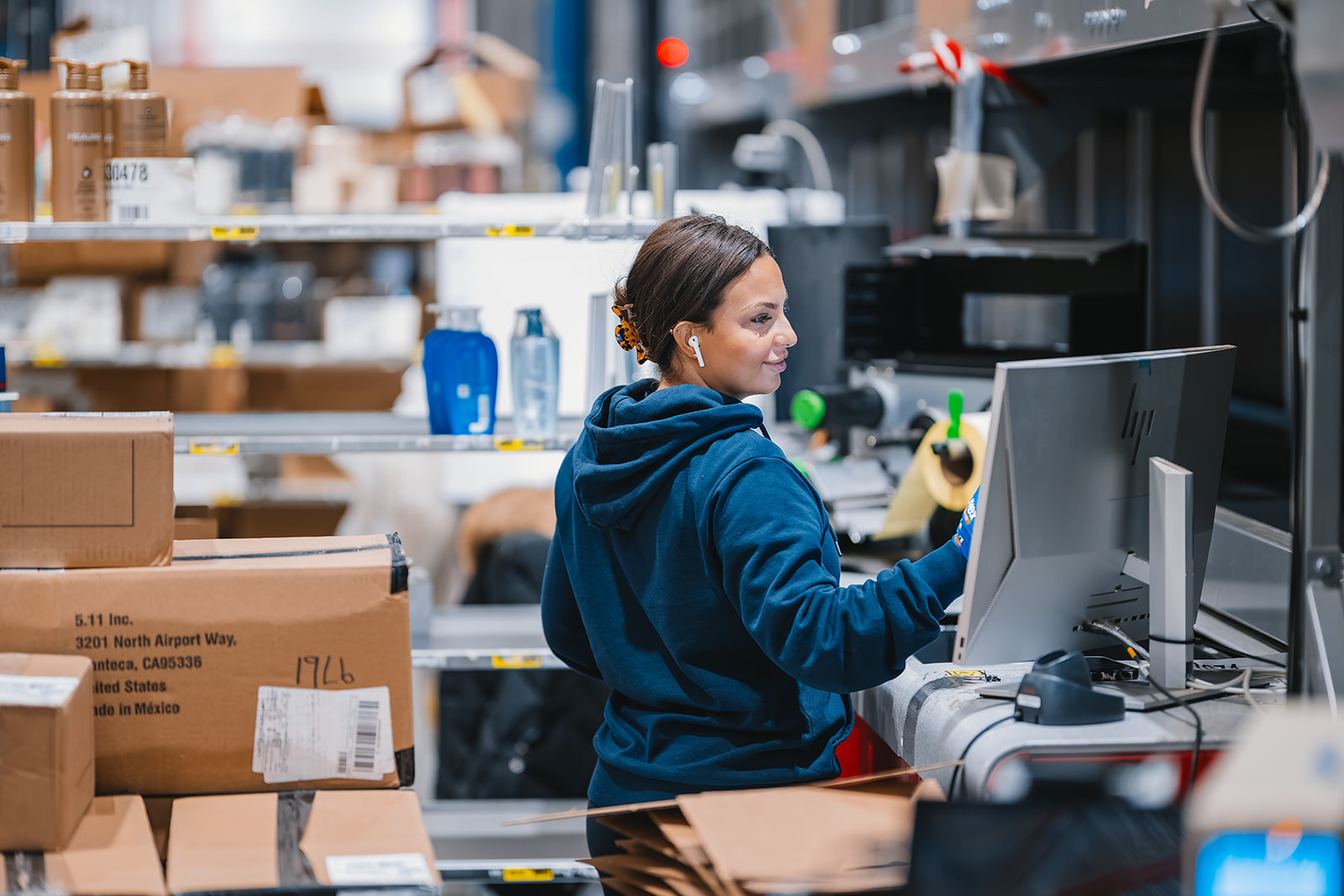 Woman in a blue hoodie working at a computer in a warehouse surrounded by stacked cardboard boxes and shelves.