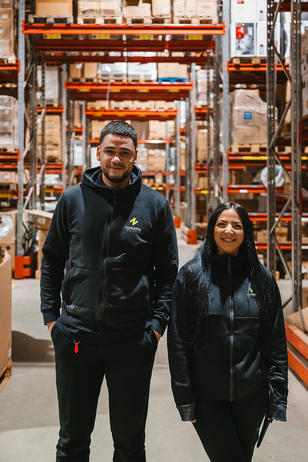 Two warehouse workers in black Nowaste Logistics jackets standing and smiling in aisle with shelves stacked with boxes.