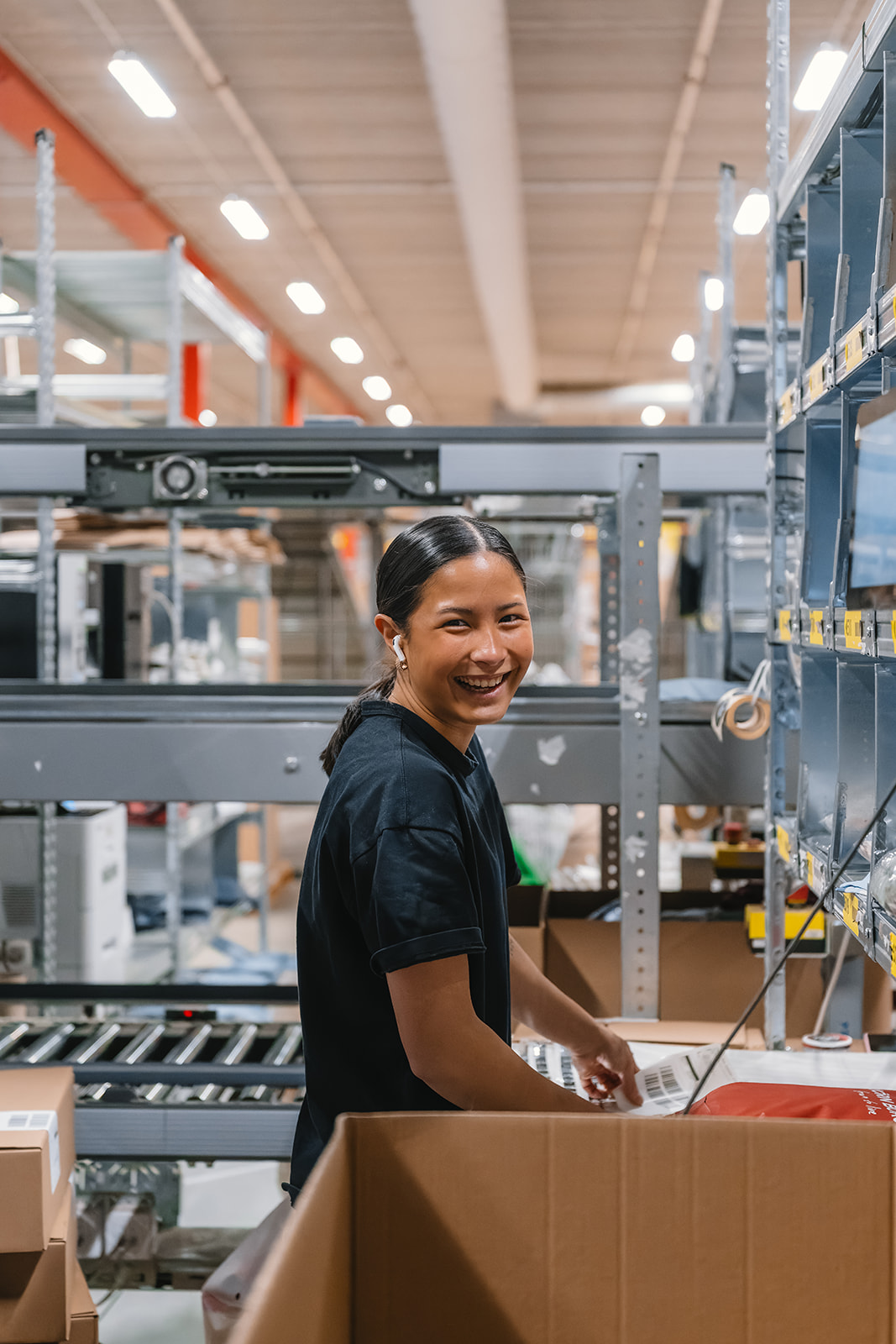 Smiling woman wearing earbuds sorting packages in a warehouse with shelves and conveyor belts.