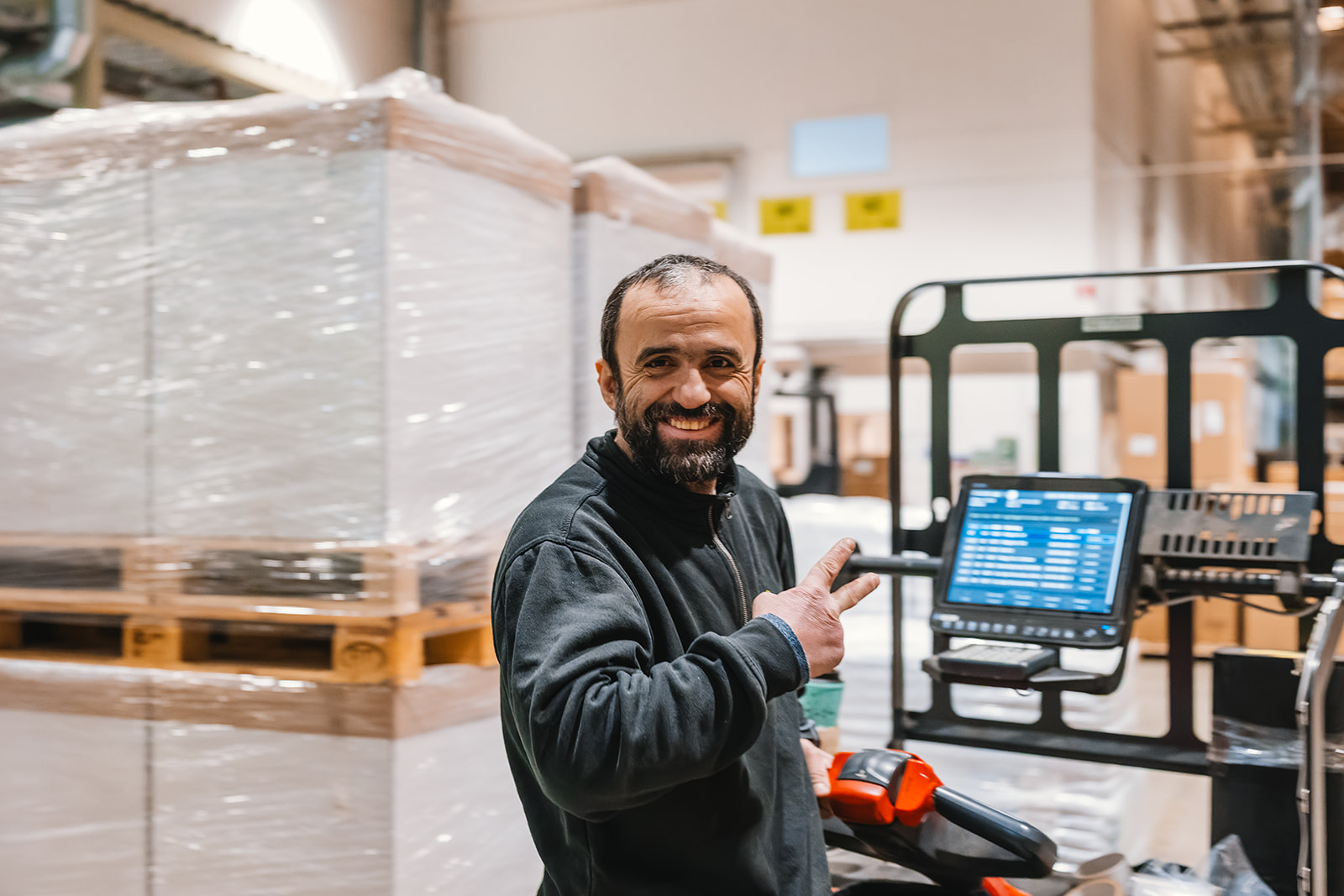 Smiling warehouse worker showing a peace sign while operating a pallet jack with a digital display in a storage area.