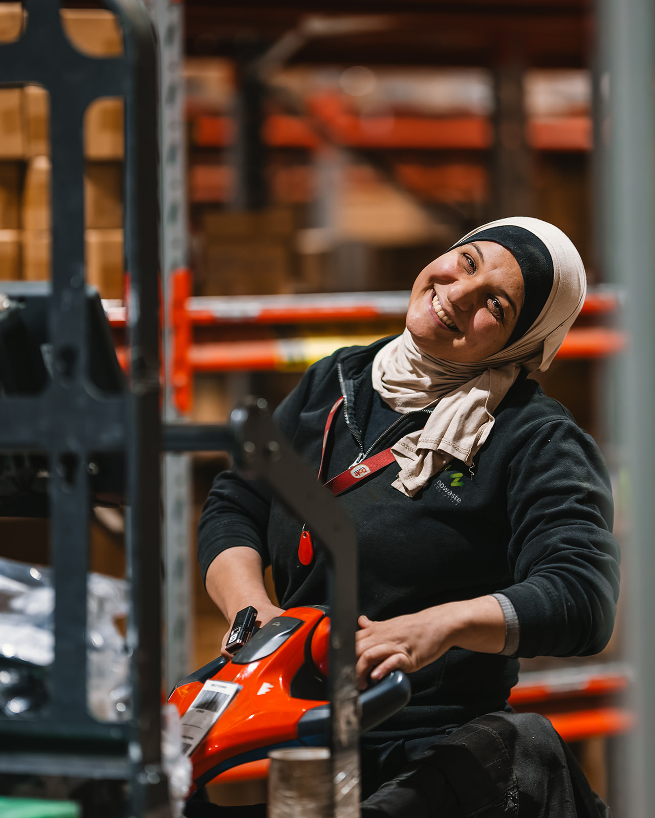 Smiling woman wearing a hijab operating a forklift in a warehouse filled with shelves and boxes.