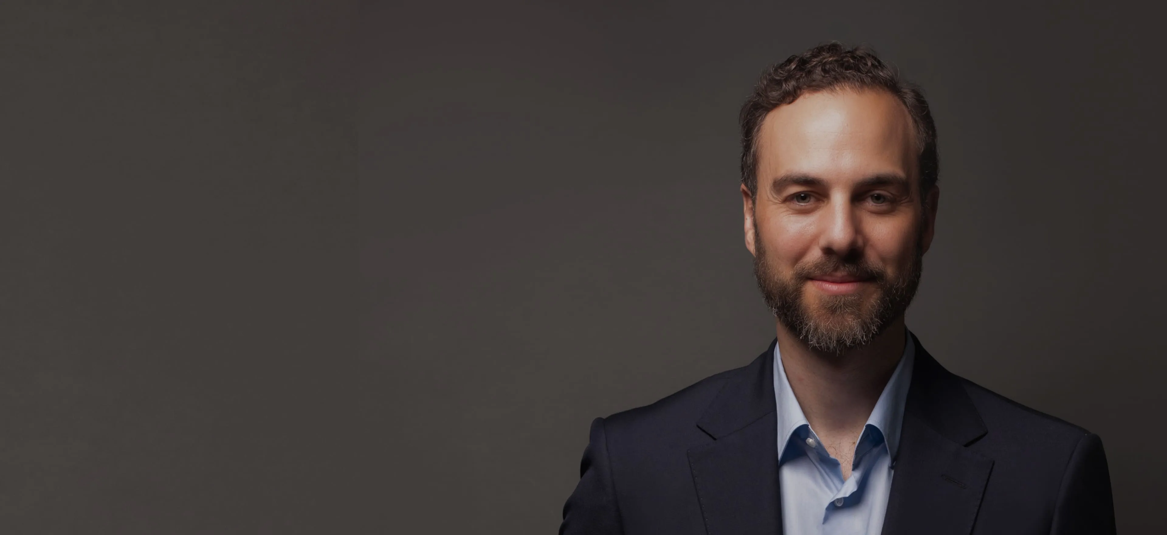 Smiling man with short curly hair and beard wearing a navy blazer and light blue shirt against a dark background.