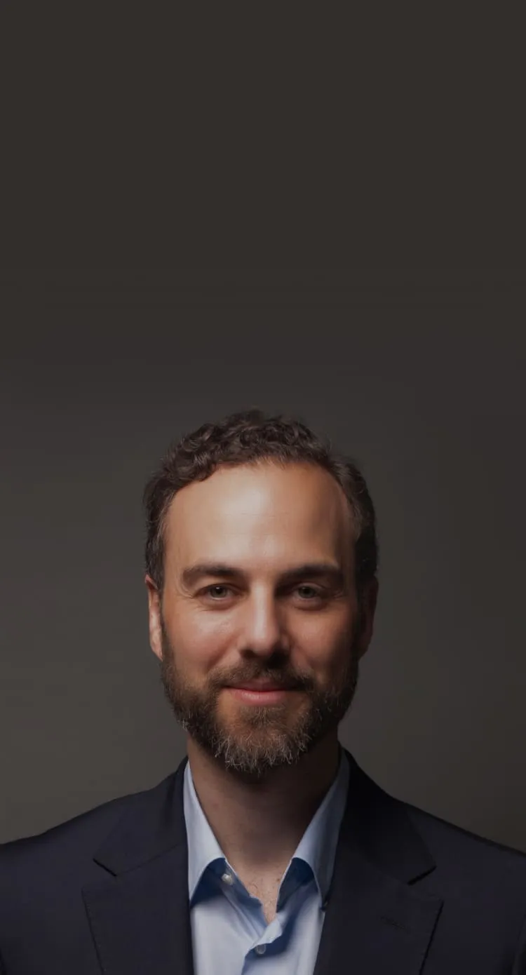 Portrait of a smiling man with curly hair and a beard wearing a dark blazer and light blue shirt against a dark background.