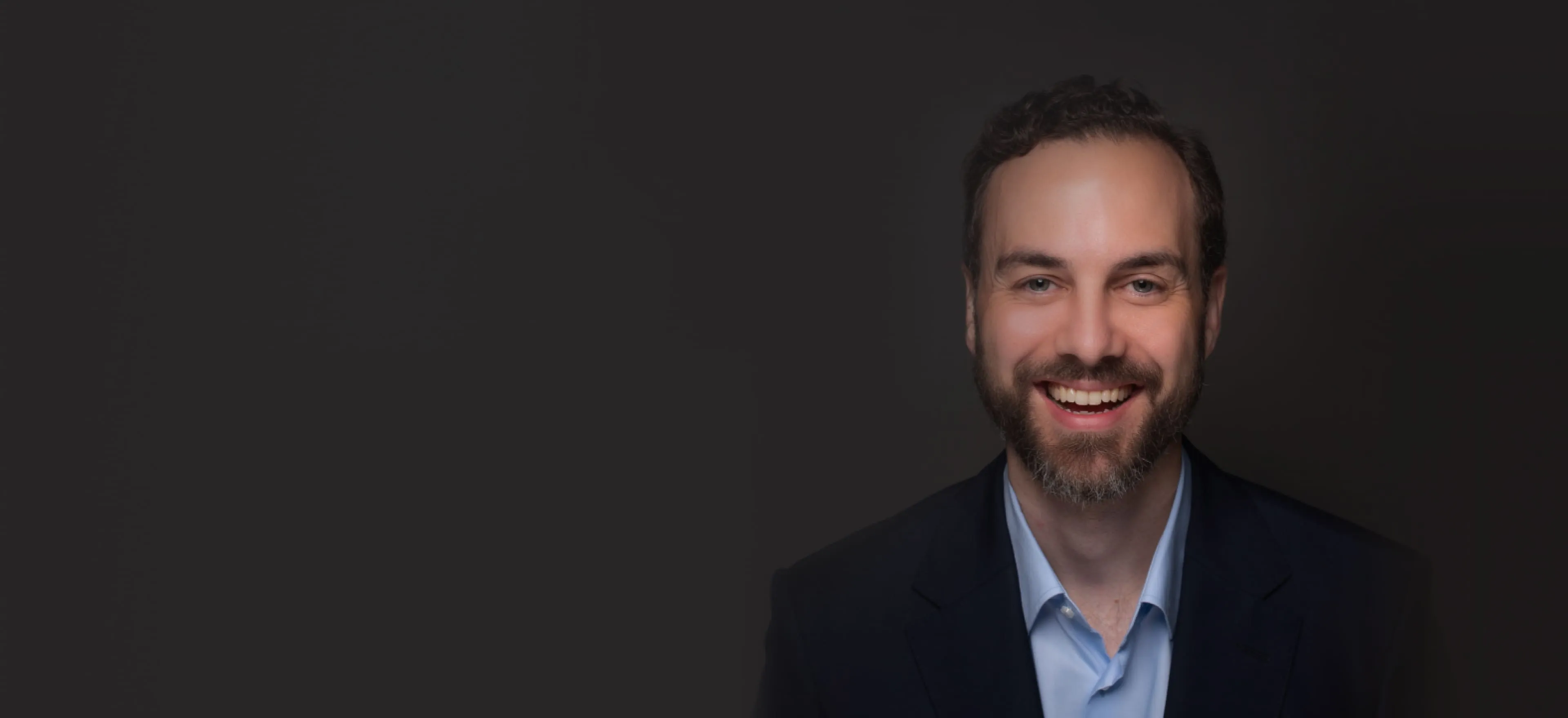 Smiling man with short curly hair and beard wearing a dark blazer and light blue shirt against a dark background.