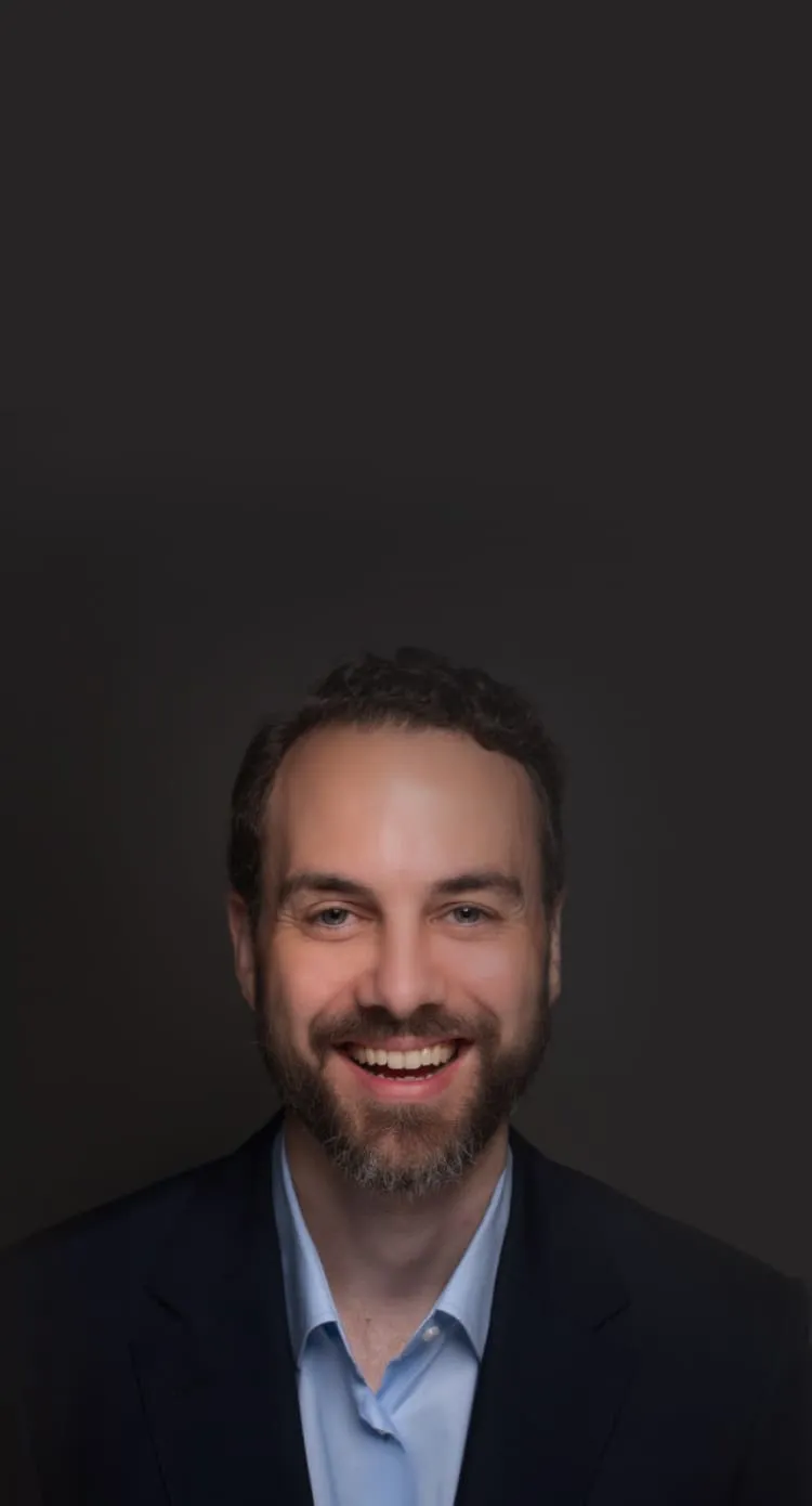 Smiling man with beard wearing a light blue shirt and dark blazer against a dark background.