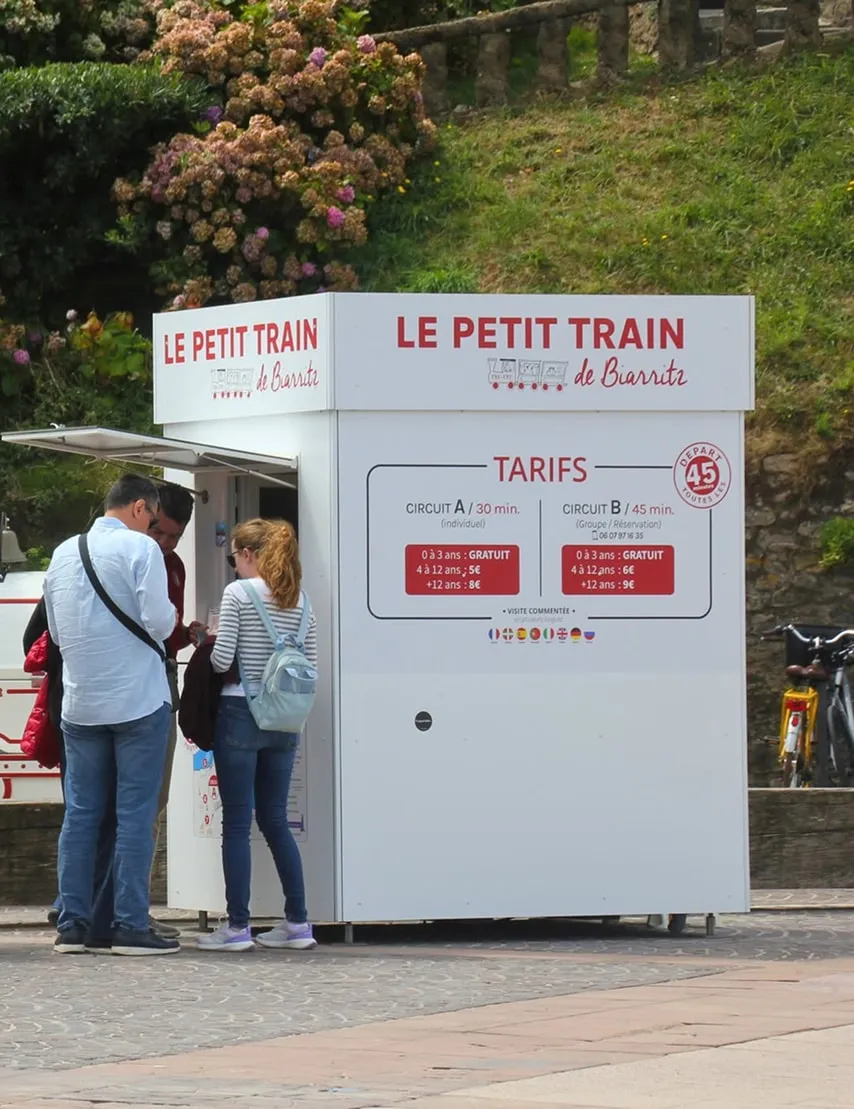 Small white ticket kiosk for Le Petit Train de Biarritz with three people standing nearby, surrounded by greenery and flowers.