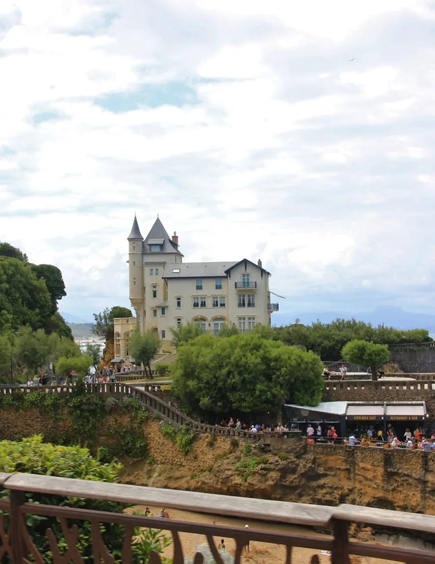 A beige castle-like building surrounded by greenery and a crowd of people near a cafe on a cliffside under a cloudy sky.