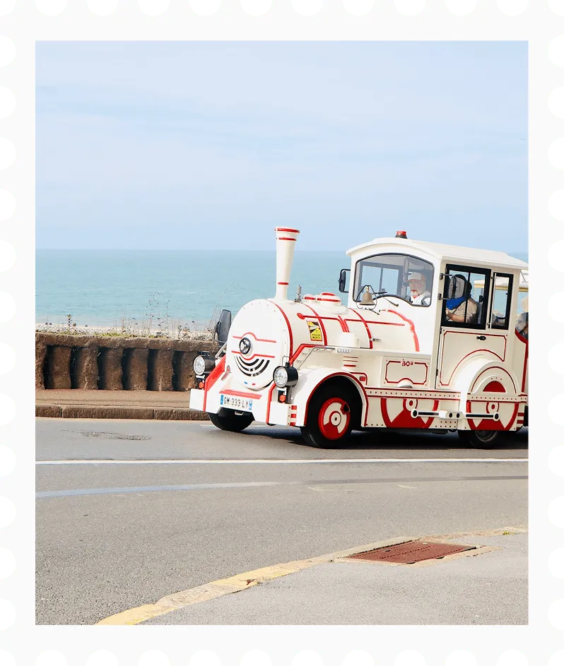 Small white and red tourist train driving along a road near a seaside promenade with the ocean in the background.
