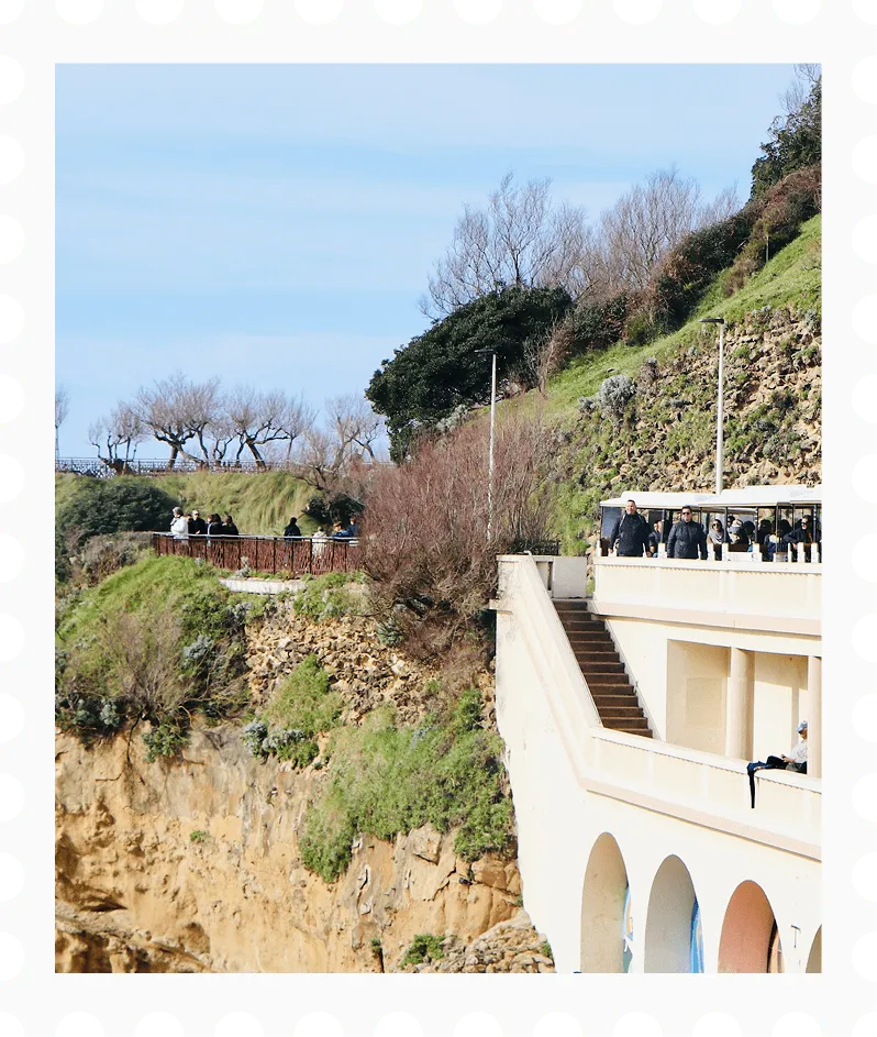 People walking and standing on a cliffside path and terrace overlooking rocky cliffs with green vegetation under a clear blue sky.