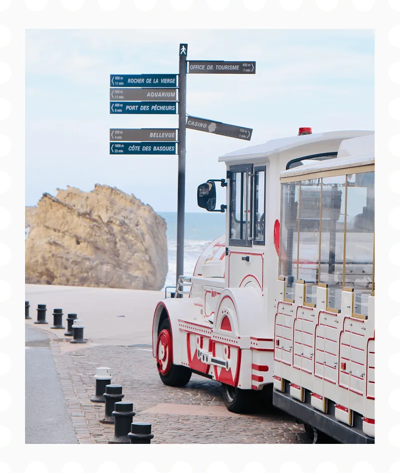 White tourist train with red accents parked near a coastal viewpoint with directional signs to local attractions and a large rock by the sea.
