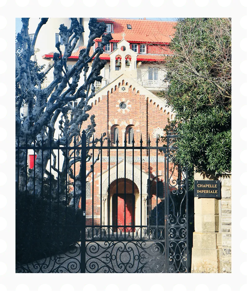 Iron gate with intricate designs in front of Chapelle Imperiale, a brick chapel with arched doorway and red roof tiles.