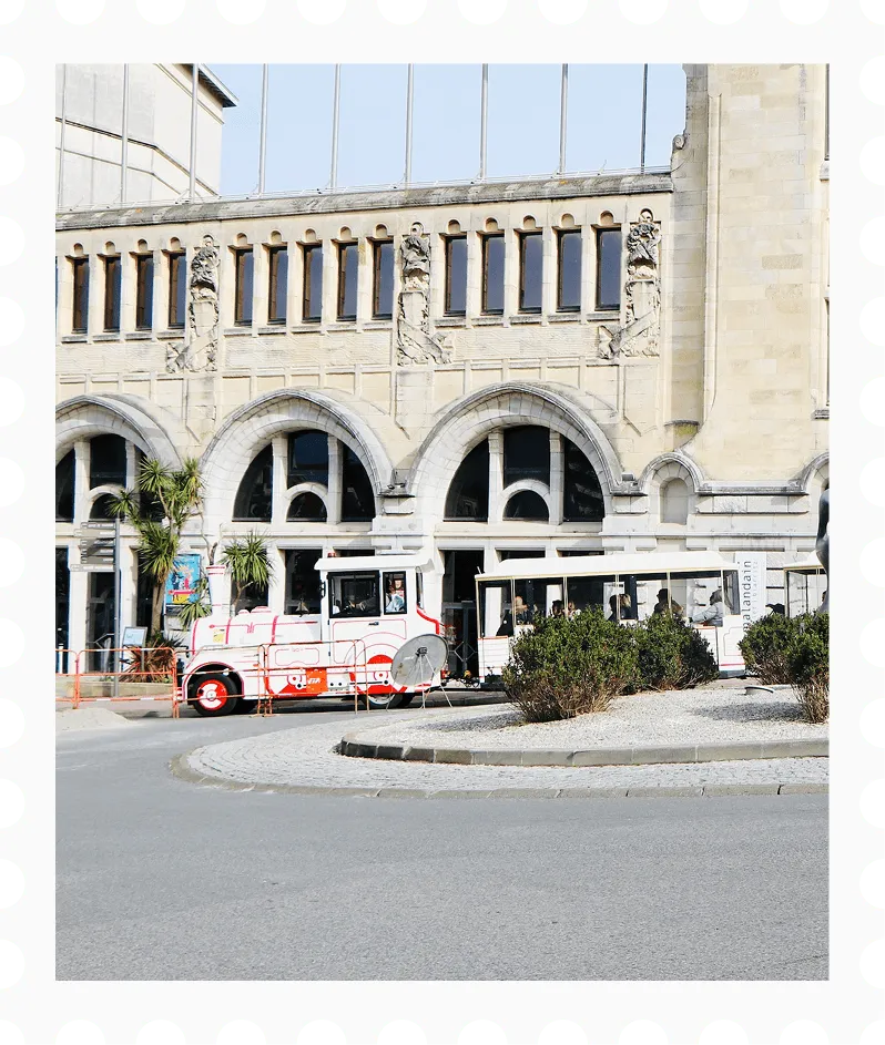 Tourist train with a white and red engine and passenger cars passing in front of a historic building with arched windows and stone sculptures.