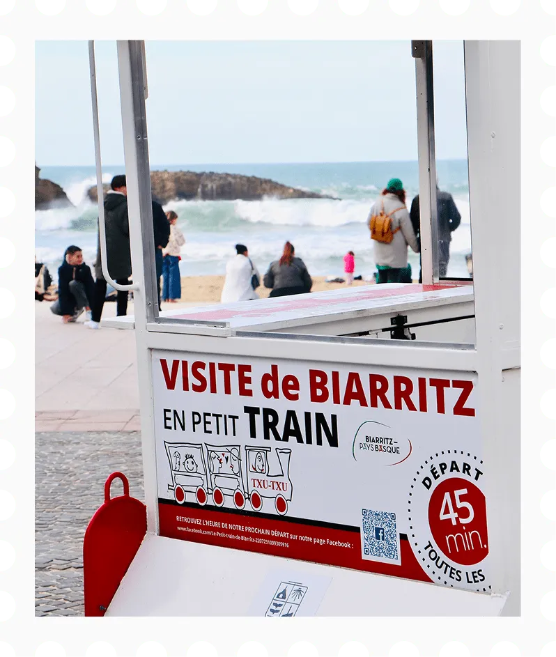 Sign advertising a small train tour in Biarritz with beach and people in the background.