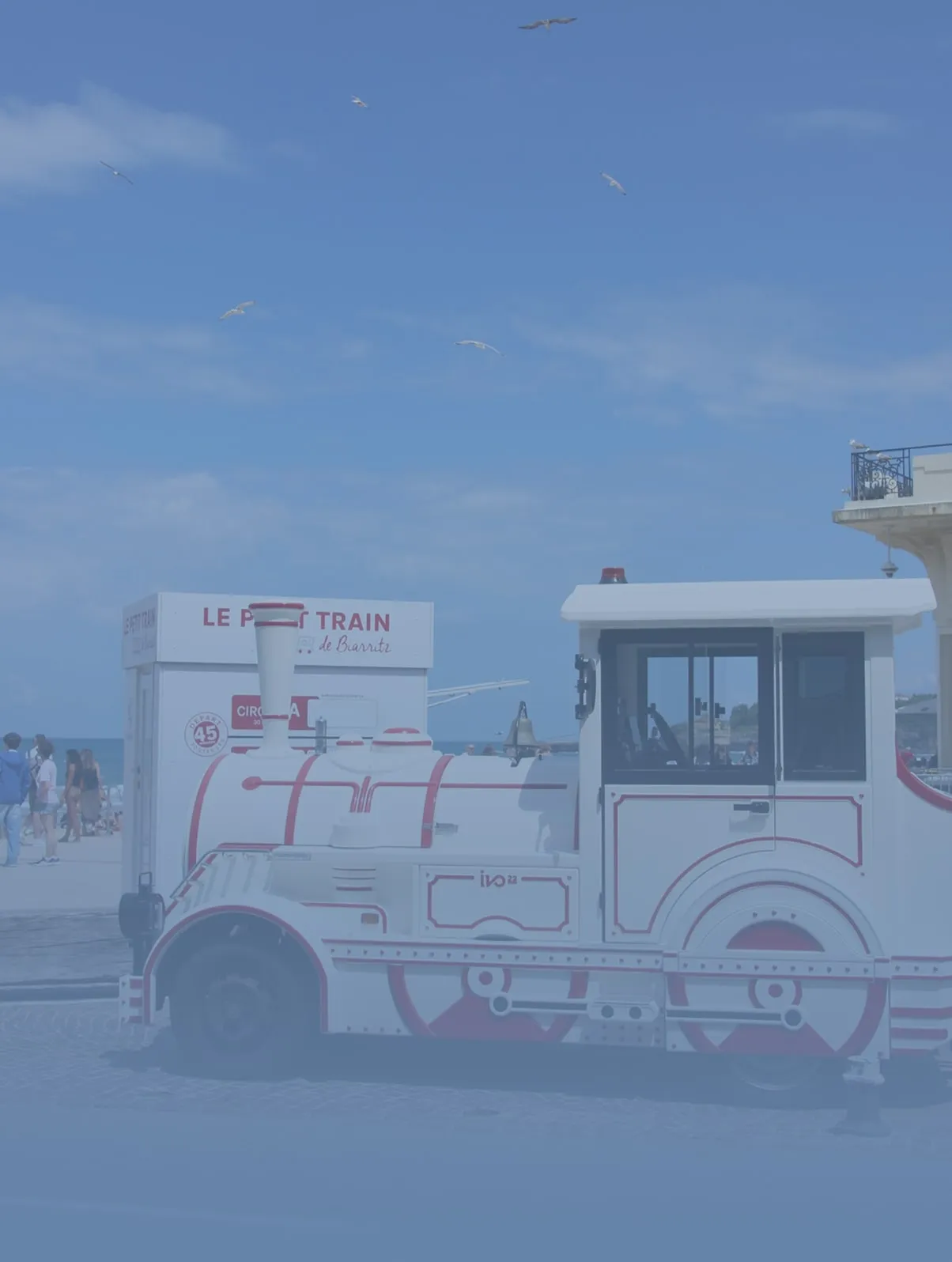 White and red miniature tourist train with people walking nearby under a blue sky with flying birds.