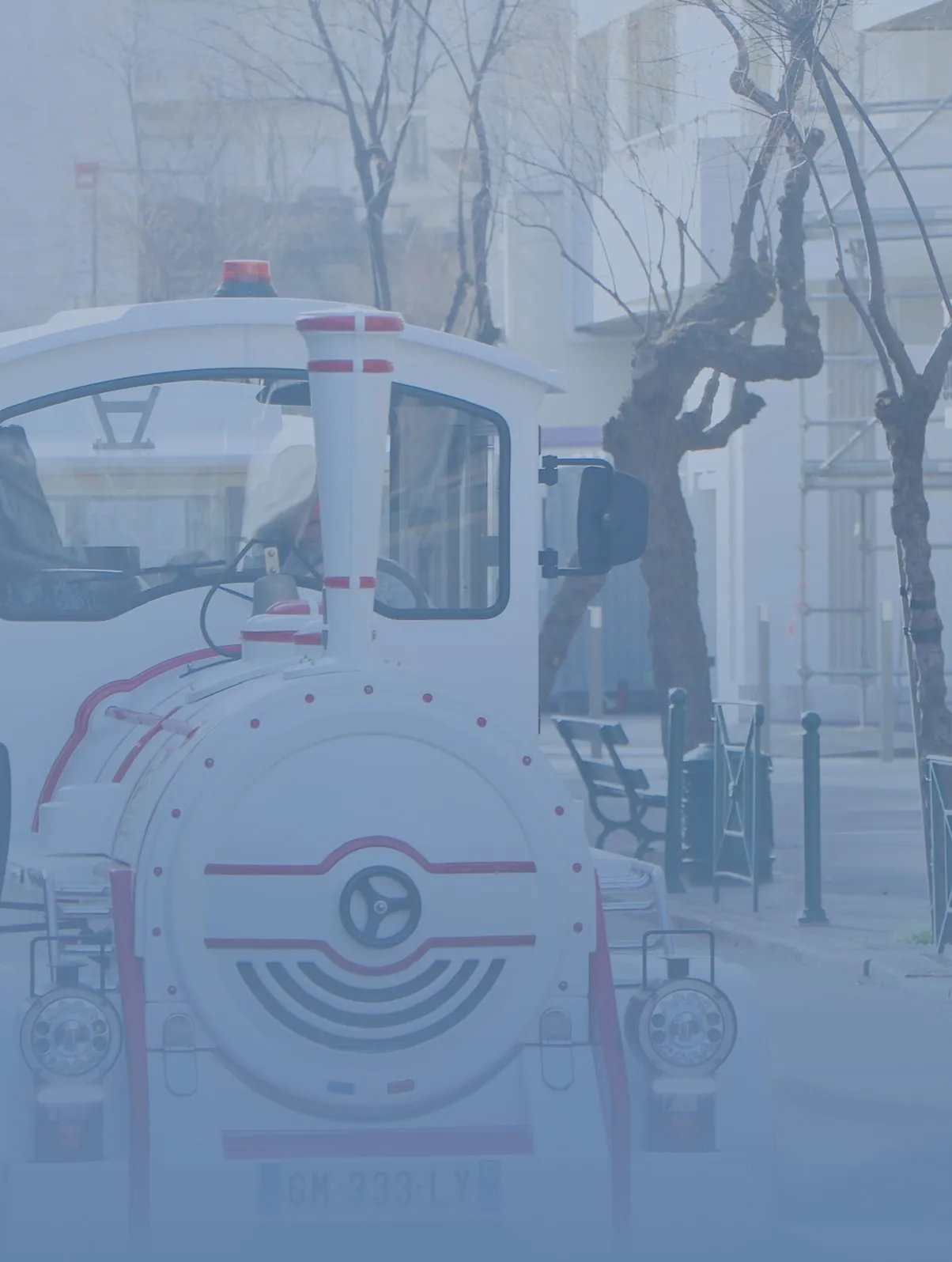 Front view of a white and red miniature street train on an urban street with leafless trees and benches.