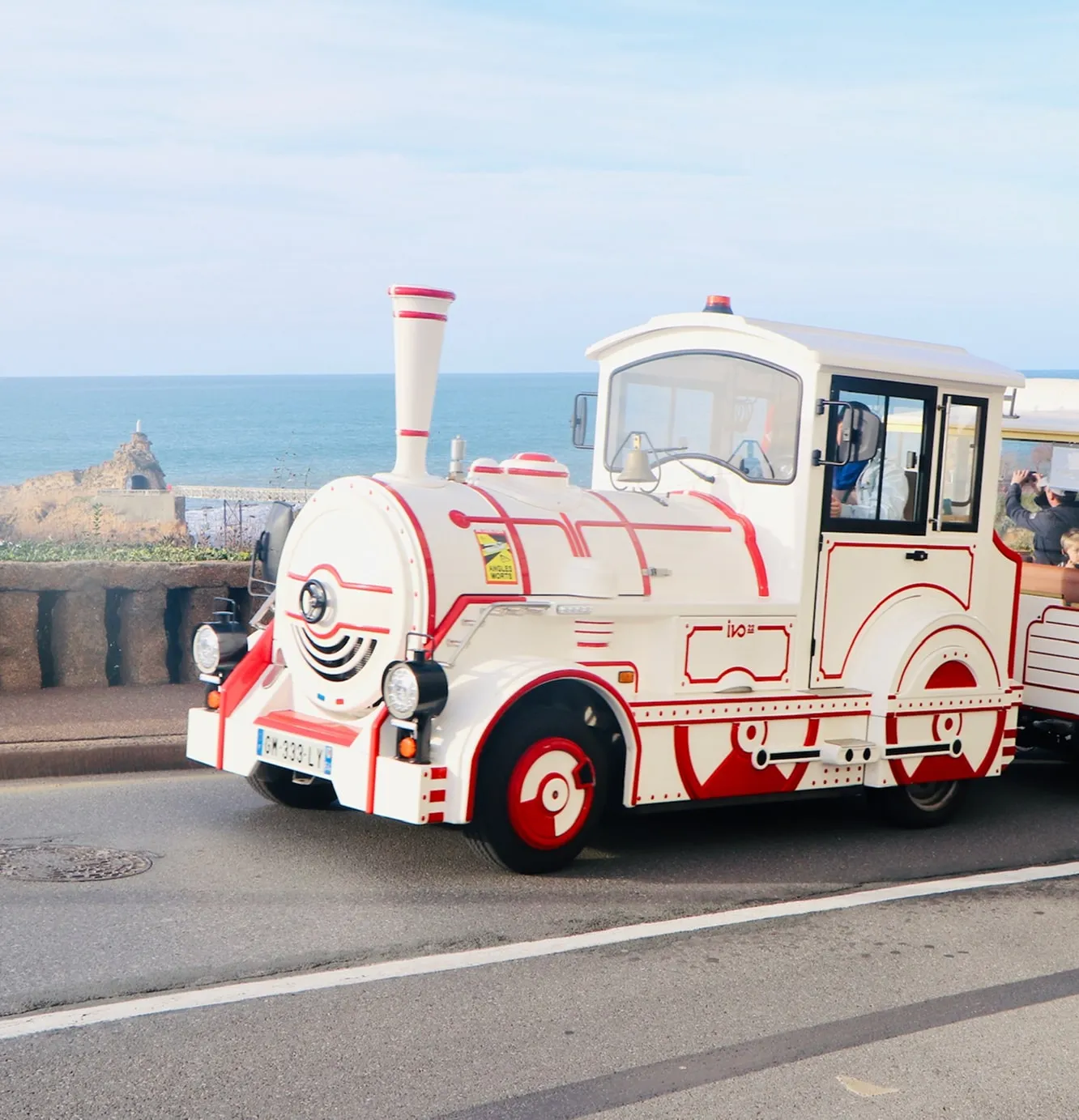 White and red tourist train with a vintage locomotive design driving along a seaside road with ocean and rocky coastline in the background.