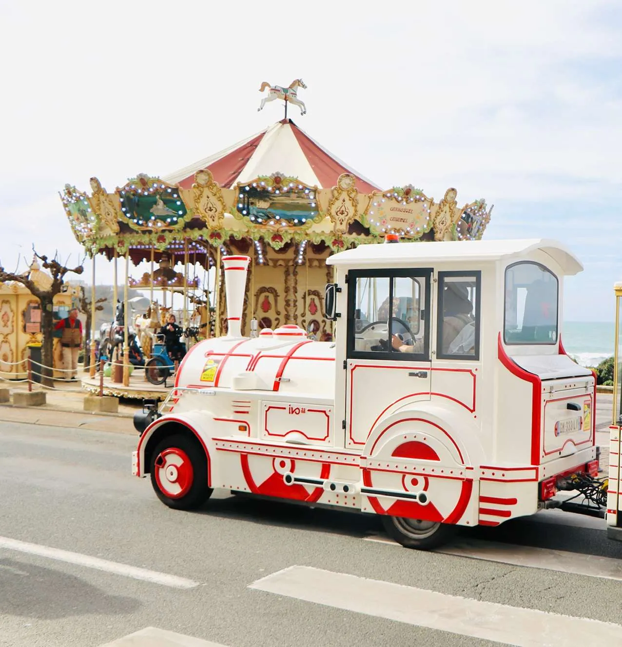White and red miniature train on a road with a vintage carousel and people in the background near the seaside.