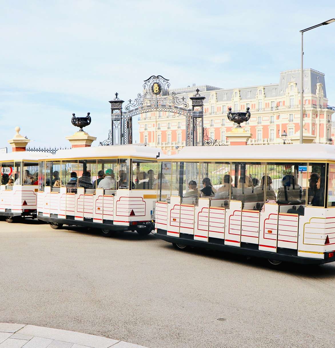 Tourist train with passengers passing by ornate iron gates and a large historic building.