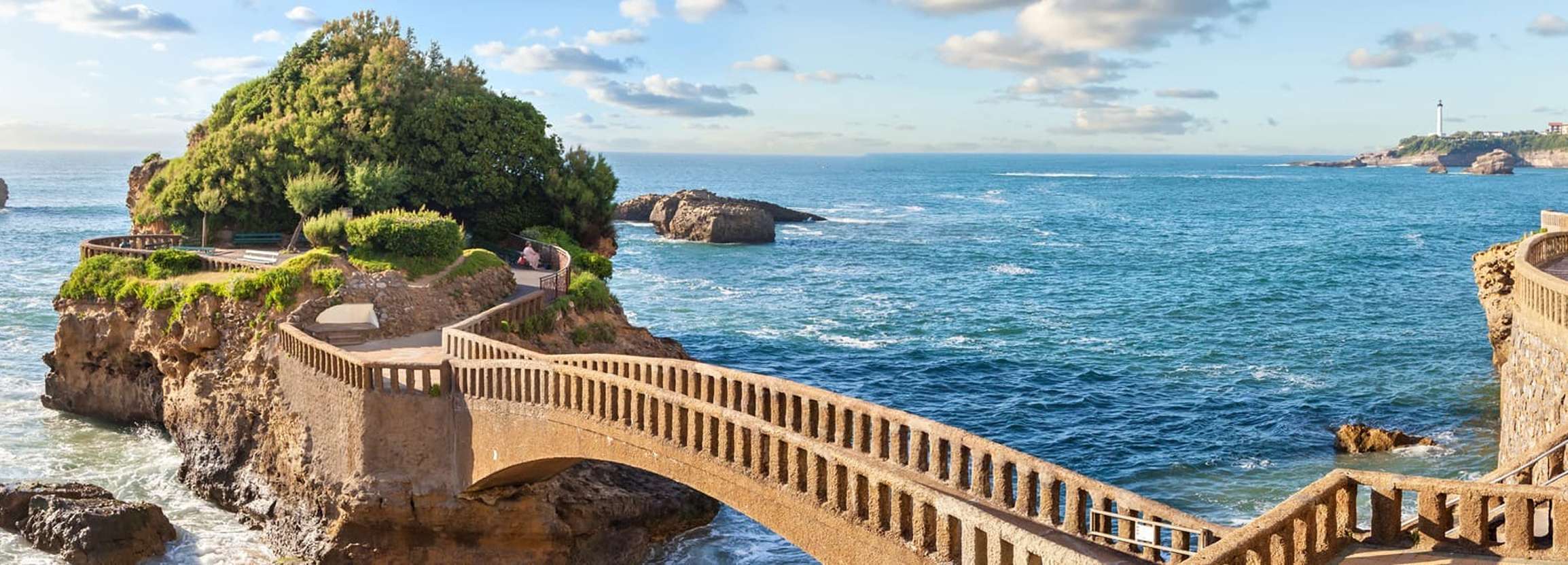 Stone bridge connecting rocky cliffs with greenery overlooking a blue ocean under a partly cloudy sky.