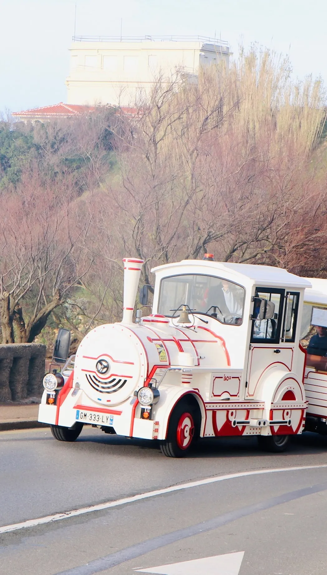 Small white and red road train driving on a curved street with leafless trees and a building in the background.