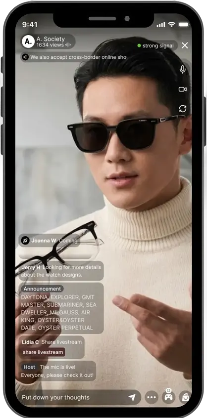 A man wearing black sunglasses and a white turtleneck shirt pointing at a pair of sunglasses he is holding during a live online video stream on a smartphone screen.