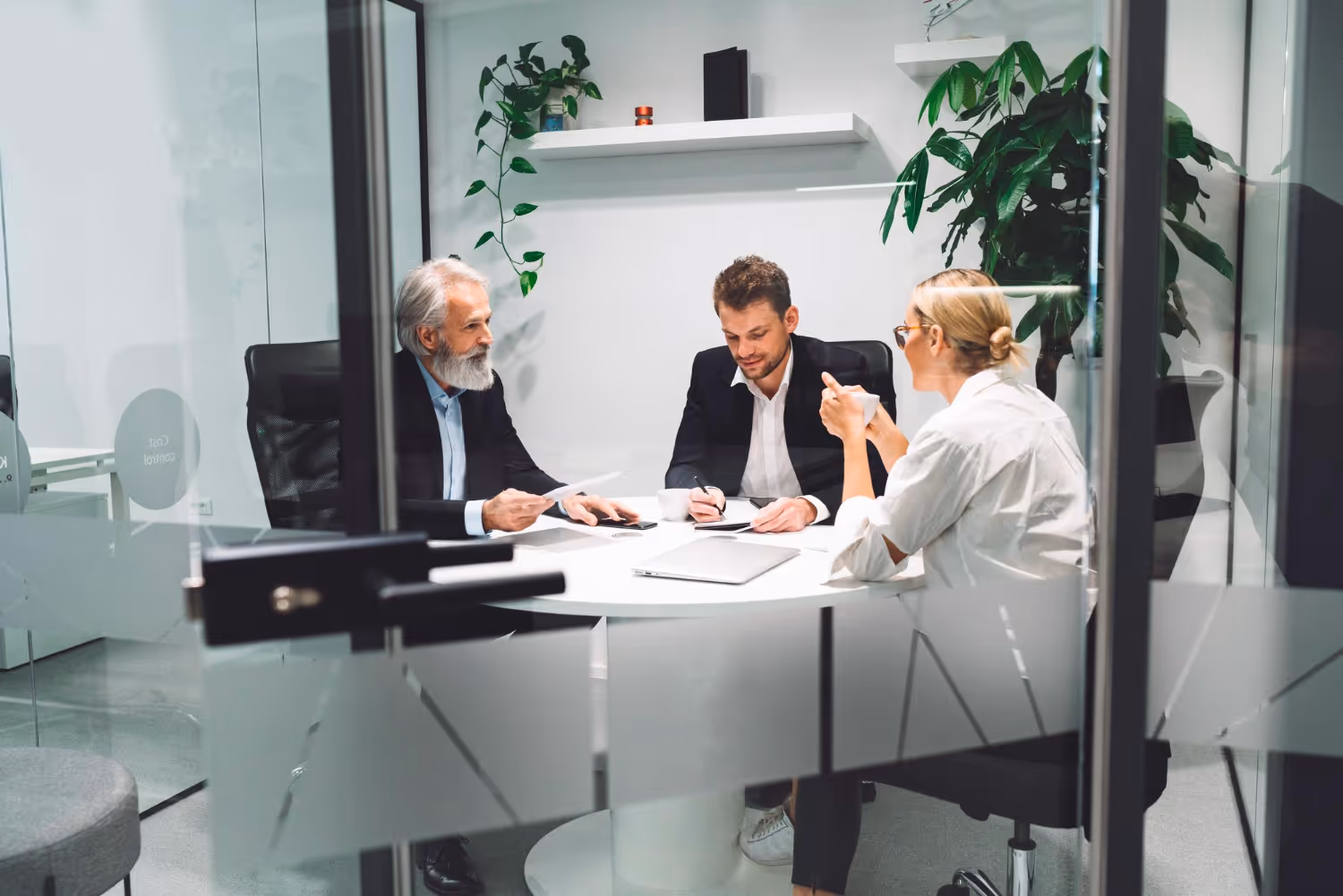 Trois professionnels en tenue de bureau tiennent une réunion autour d’une table ronde blanche dans un bureau moderne avec des plantes.